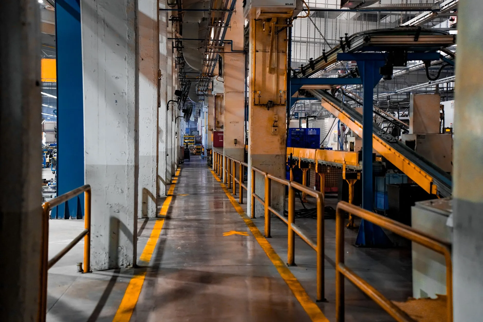 Walkway marked with yellow lines in a factory, lined with guardrails and an industrial conveyor