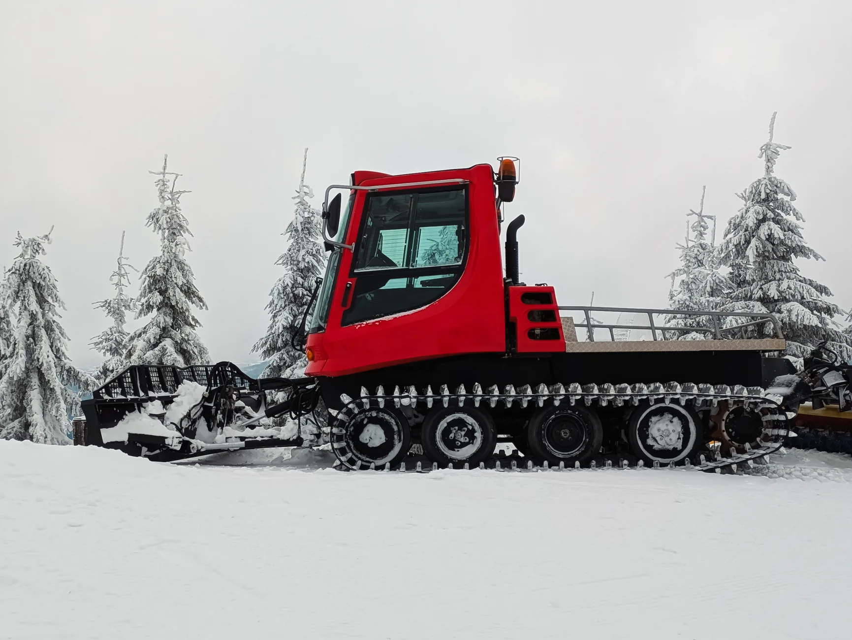 Red tracked vehicle with a front blade parked on a snowy trail lined with fir trees
