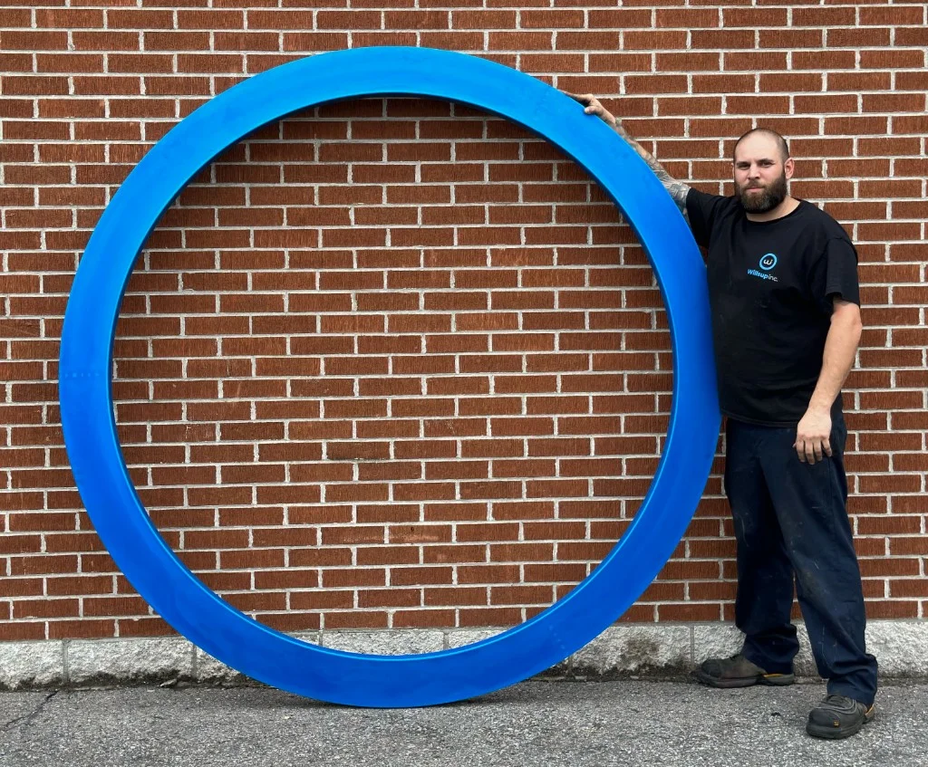 Membre de l’équipe Willsup en chandail de l’entreprise, à côté d’un grand anneau industriel bleu en polyuréthane devant un mur de brique rouge
