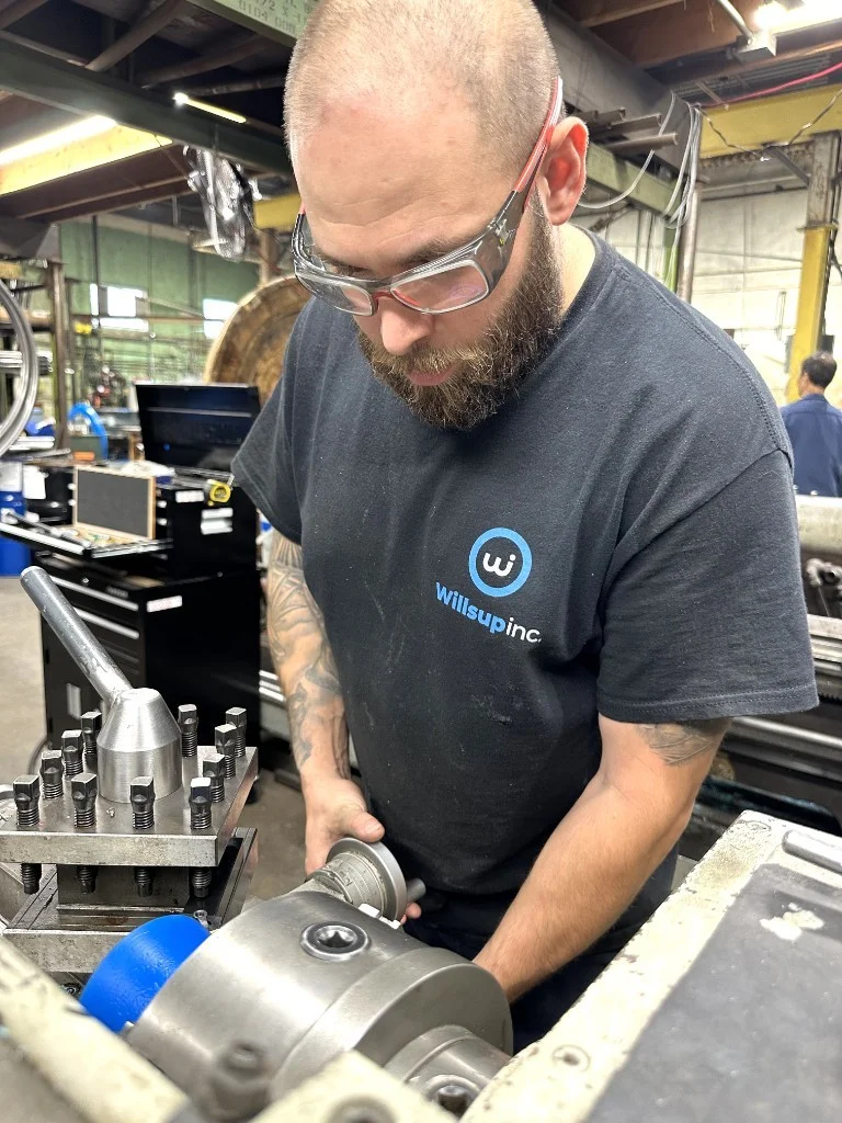 Willsup machinist with beard and safety glasses operating a metal lathe on a blue cylindrical part in an industrial workshop